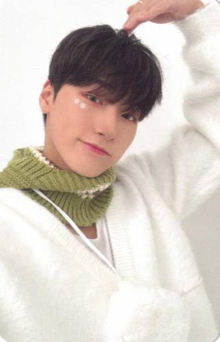 Photocard of San wearing a white sweater and green scarf, making a half heart with his arm over his head