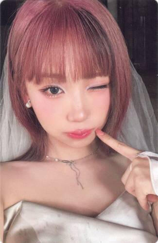 Photocard of Dosie wearing a wedding dress, poking her cheek, and winking
