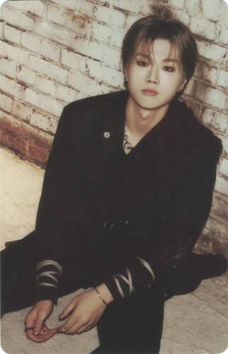 Photocard of Jungsu wearing a black outfit, sitting against a brick wall, and looking up at the camera