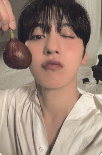 Photocard of Keonhee wearing a white button up and winking at the camera, holding a fig by his face
