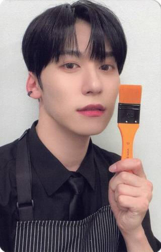 Photocard of Keonhee wearing a black button up and smock, holding an orange paint brush