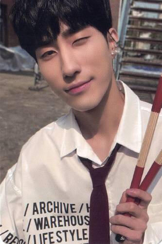 Photocard of Harin wearing a white shirt with red tie, holding a pair of drumsticks, and winking