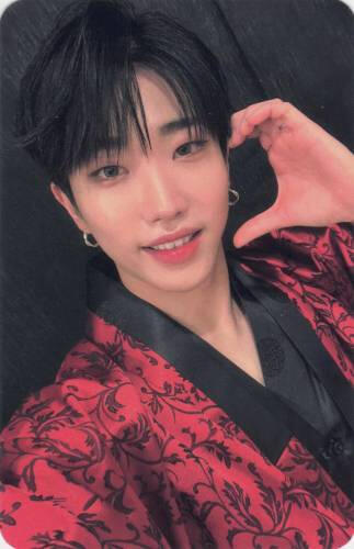 A photocard of Dann in red floral hanbok, making a half heart on his fake and toothily grinning