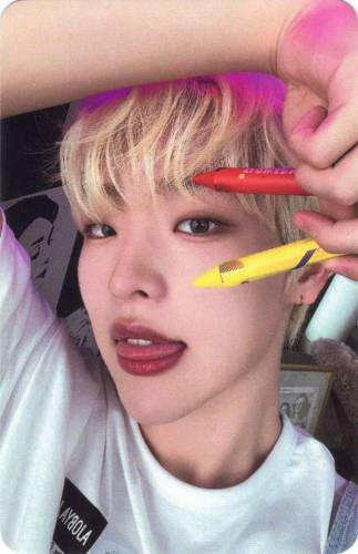 Photocard of Mujin holding two crayons by his eye and sticking his tongue out